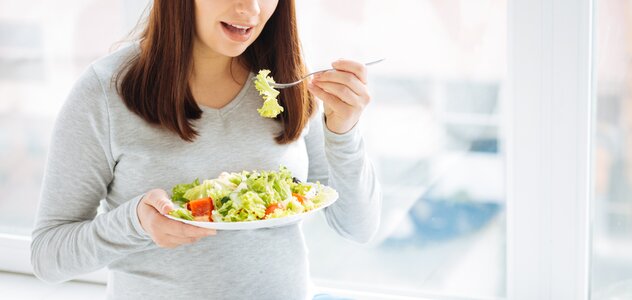 Kobieta w ciąży siedzi na parapecie z talerzem pełnym zdrowej żywości. Przyszła mama zjada widelcem zdrową sałatkę z warzywami. 