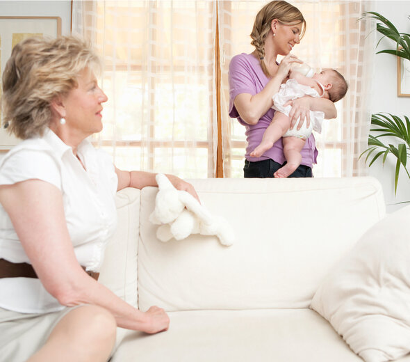 Babcia przyszła w odwiedziny zobaczyć swojego nowonarodzonego wnuczka. Kobieta z dzieckiem stoi za kanapą, a babcia przygląda się maluszkowi. 