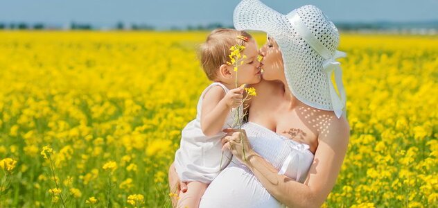 Młoda kobieta w ciąży stoi na polu żółtych kwiatów, na rękach trzyma dziecko. Ma słomkowy kapelusz i ubrana jest na biało. 