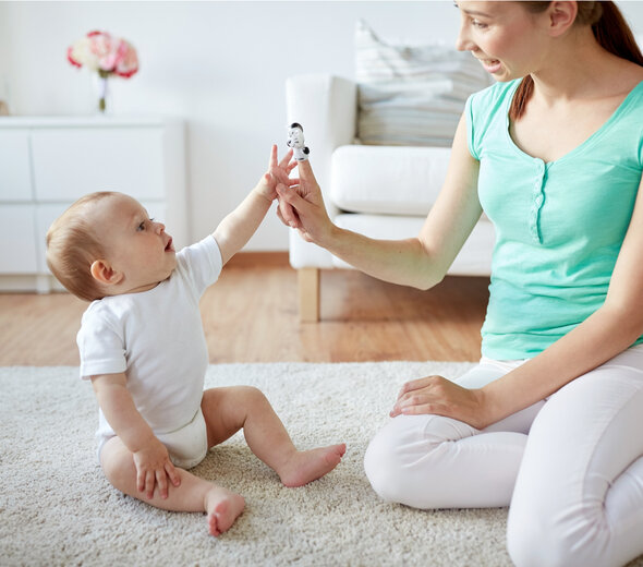 Mama bawi się z dzieckiem na dywanie. Bawią się palcami, mama pokazuje szmacianą zabawkę na jej palcu. Za nimi stoi biała kanapa i biały regał. Mama jest ubrana w bluzkę w kolorze morskim i białe spodnie, a dziecko w białe body. 