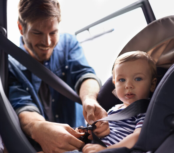 Tata zapina dziecko w foteliku samochodowym. Ma blond włosy i dżinsową koszulę. Niemowlę patrzy w obiektyw. Ma na sobie granatowo białą koszulkę w paski, z krótkim rękawem. 