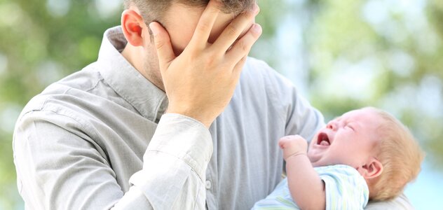 Tata trzyma niemowlę w ramionach. Ma na sobie szarą koszulę. Pochyla głowę i zasłania dłonią twarz. Niemowlę płacze, ma na sobie błękitną koszulkę z krótkim rękawem. 