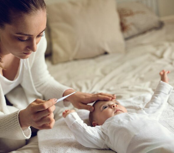 Mama siedzi na łóżku, obok leży niemowlę. Mama sprawdza dłonią czoło dziecka, w drugiej dłoni trzyma termometr. Patrzy na termometr. Ma na sobie biały sweter. Dziecko jest również ubrane na biało. 