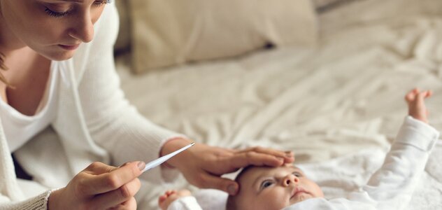 Mama siedzi na łóżku, obok leży niemowlę. Mama sprawdza dłonią czoło dziecka, w drugiej dłoni trzyma termometr. Patrzy na termometr. Ma na sobie biały sweter. Dziecko jest również ubrane na biało. 