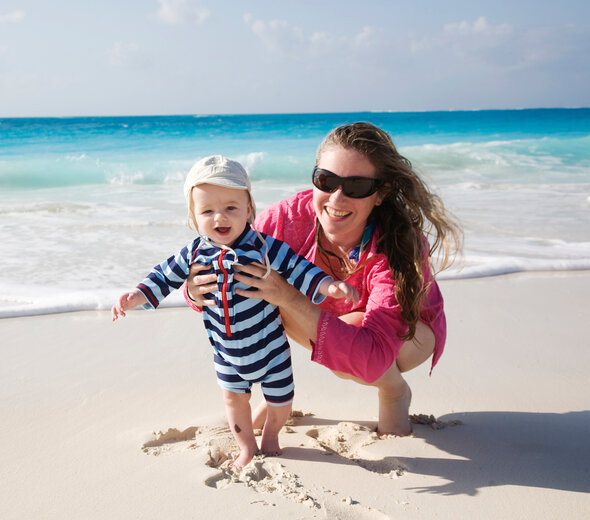 Mama i dziecko są na plaży nad morzem. Mama kuca na piasku i podtrzymuje małe dziecko. Ma długie brązowe włosy, okulary przeciwsłoneczne i różową bluzkę.  Dziecko ma czapeczkę z daszkiem na głowie. Jest ubrane w biało granatowy  rampers w paski. 