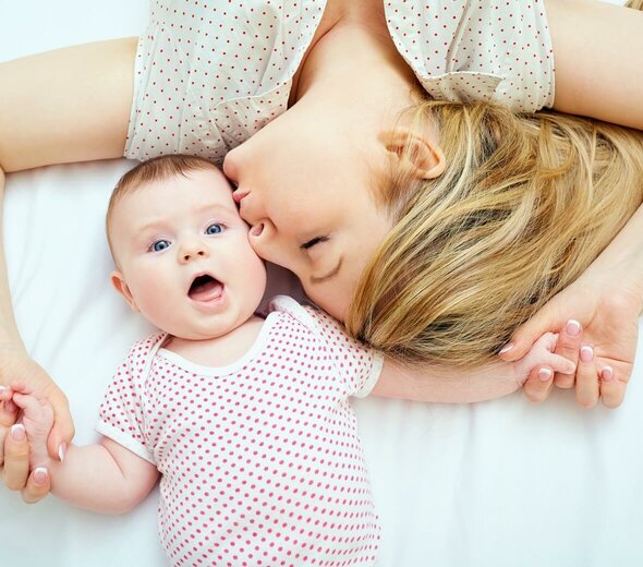 Mama leży z niemowlakiem na łóżku na białym prześcieradle. Trzyma niemowlę za rączki i całuje je w główkę. Oboje mają na sobie ubrania w kropki. 