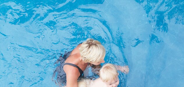 Mama podtrzymuje rękami niemowlę w basenie. Mama i niemowlę mają krótkie blond włosy. Mama ma na sobie granatowy kostium jednoczęściowy. 