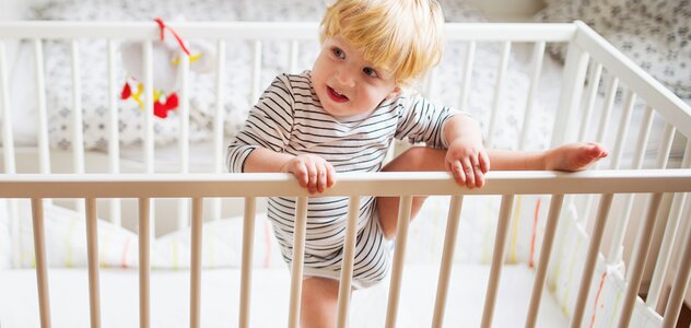 Dziecko wspina się na bok łóżeczka ze szczebelkami. Próbuje wyjść z łóżeczka. Ma blond włosy i białe body w paski. W tle widać łóżko rodziców. 