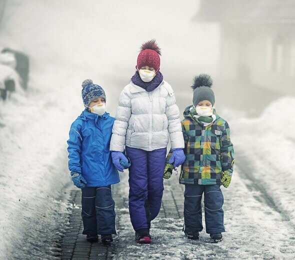 Mama i dwoje dzieci idą zaśnieżonym chodnikiem. Mają na sobie kombinezony zimowe, a na twarzy maski antysmogowe. Na głowie noszą czapki. Mama ma białą kurtkę i fioletowe spodnie. Dziecko ma niebieską kurtkę i granatowe spodnie. Drugie dziecko ma kolorową kurtkę i granatowe spodnie. 