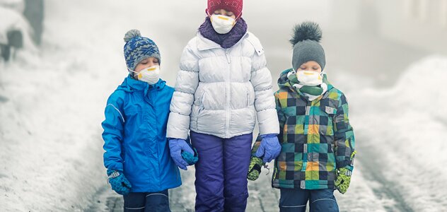 Mama i dwoje dzieci idą zaśnieżonym chodnikiem. Mają na sobie kombinezony zimowe, a na twarzy maski antysmogowe. Na głowie noszą czapki. Mama ma białą kurtkę i fioletowe spodnie. Dziecko ma niebieską kurtkę i granatowe spodnie. Drugie dziecko ma kolorową kurtkę i granatowe spodnie. 