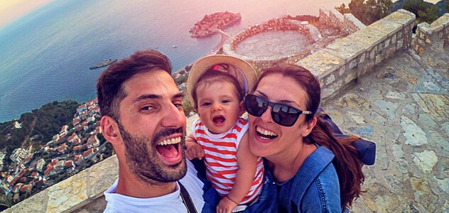 Mama, tata i dziecko stoją na chodniku i robią sobie selfie. Uśmiechają się do obiektywu. Tata trzyma dziecko na rękach. Dziecko ma na głowie kapelusik. Mama ma okulary przeciwsłoneczne.  W tle na dole widać miasteczko i morze. 