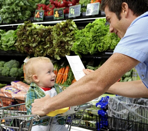 Tata i dziecko są w supermarkecie, przy półce z warzywami. Dziecko siedzi w koszyku na zakupy. Tata trzyma kartkę z listą rzeczy do kupienia. Dziecko patrzy na tatę i się uśmiecha. 