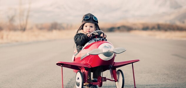 Dziecko siedzi w rowerku o kształcie samolotu w kolorze czerwonym. Ma czapkę pilotkę i lotnicze okulary. W tle widać ulicę i góry. 