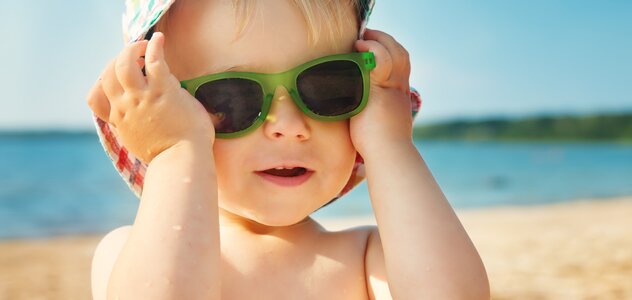 Dziecko jest na plaży. W tle widać morze. Dziecko ma na głowie materiałowy kapelusik a na oczach zielone okulary słoneczne, które podtrzymuje rączkami. 