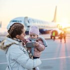 Mama stoi na płycie lotniczej i  trzyma na rękach córkę. W tle widać samolot. Mama ma długie włosy zaplecione w warkocze i białą kurtkę puchową. Córka ma różową kurtkę i różową czapkę. 