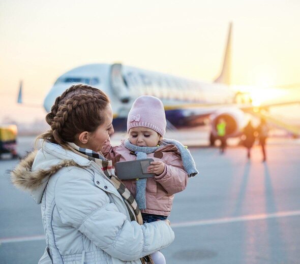 Mama stoi na płycie lotniczej i  trzyma na rękach córkę. W tle widać samolot. Mama ma długie włosy zaplecione w warkocze i białą kurtkę puchową. Córka ma różową kurtkę i różową czapkę. 