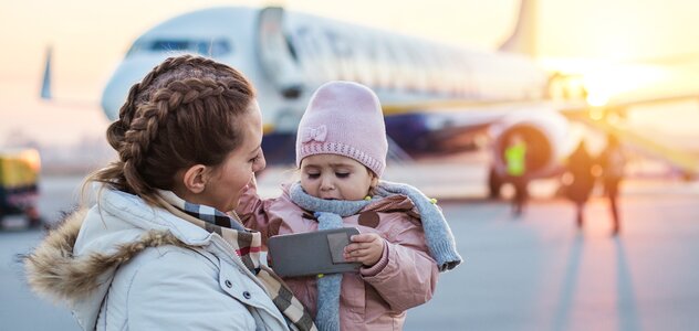 Mama stoi na płycie lotniczej i  trzyma na rękach córkę. W tle widać samolot. Mama ma długie włosy zaplecione w warkocze i białą kurtkę puchową. Córka ma różową kurtkę i różową czapkę. 