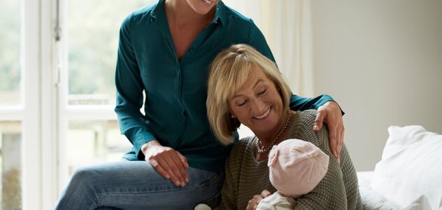 Babcia siedzi na kanapie i trzyma w objęciach niemowlę. Uśmiecha się do niego. Na brzegu kanapy siedzi mama. Uśmiecha się do niemowlęcia. Na kanapie leżą dwie białe poduszki. W tle widać okno. 