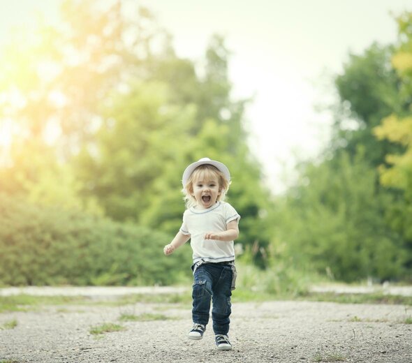 Dziecko biegnie po chodniku w parku. Patrzy w obiektyw i się śmieje. Ma blond włosy, kapelusik, białą koszulkę, dżinsy i trampki. W tle widać zieleń. 