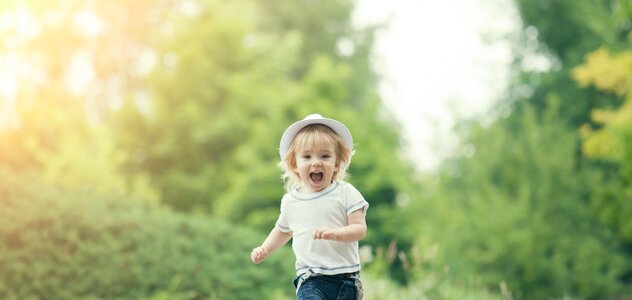 Dziecko biegnie po chodniku w parku. Patrzy w obiektyw i się śmieje. Ma blond włosy, kapelusik, białą koszulkę, dżinsy i trampki. W tle widać zieleń. 