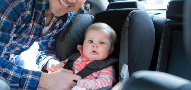 Tata przypina niemowlę w aucie w foteliku samochodowym. Tata i niemowlę patrzą w obiektyw. Tata ma na sobie niebieską koszulę w kratę. Niemowlę ma różowo szarą bluzkę w paski. 