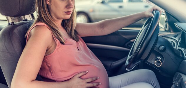 Kobieta w ciąży siedzi na miejscu kierowcy w aucie. Jedną ręką trzyma na kierownicy, drugą dotyka brzucha. Ma długie blond włosy, różową bluzkę na ramiączkach i białe spodnie. Patrzy przed siebie. Okno w samochodzie jest uchylone. 