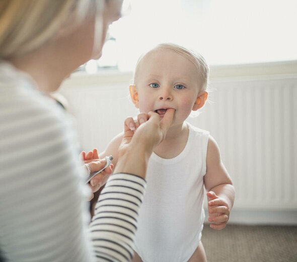 Mama kuca przy dziecku. Jedną ręką smaruje mu maścią ząb, w drugiej trzyma maść. Dziecko jest ubrane w białe body. Mama ma blond włosy i białą bluzkę w czarne paski. W tle widać biały kaloryfer. 