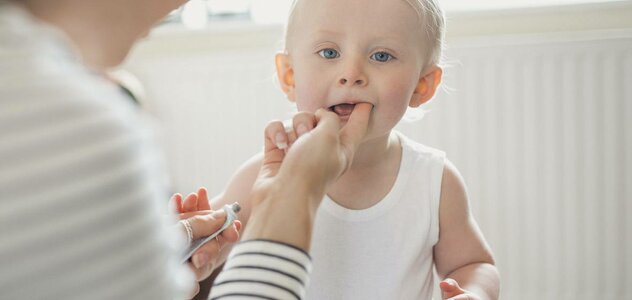 Mama kuca przy dziecku. Jedną ręką smaruje mu maścią ząb, w drugiej trzyma maść. Dziecko jest ubrane w białe body. Mama ma blond włosy i białą bluzkę w czarne paski. W tle widać biały kaloryfer. 