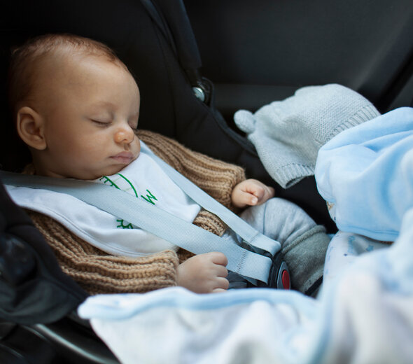 Niemowlę śpi w foteliku samochodowym w aucie. Ma beżowy sweterek. Obok leży błękitna czapeczka. Widać błękitny kocyk.