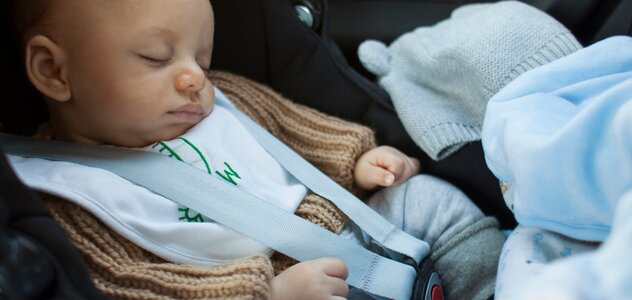 Niemowlę śpi w foteliku samochodowym w aucie. Ma beżowy sweterek. Obok leży błękitna czapeczka. Widać błękitny kocyk.