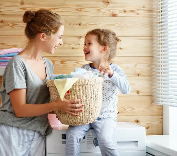 Dziewczynka i mama są w łazience. Dziewczynka siedzi na pralce. Mama i dziewczynka wspólnie trzymają koszyk do prania. Uśmiechają się do siebie. Obie mają blond włosy. Dziewczynka jest ubrana w piżamkę. Mama ma szarą koszulkę z krótkim rękawem i spodnie od piżamy. W tle widać ręczniki i drewnianą ścianę. 