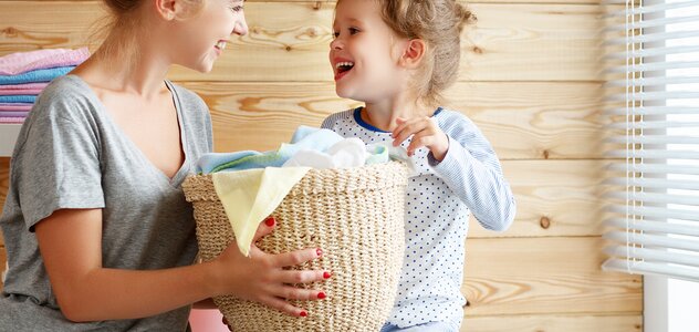 Dziewczynka i mama są w łazience. Dziewczynka siedzi na pralce. Mama i dziewczynka wspólnie trzymają koszyk do prania. Uśmiechają się do siebie. Obie mają blond włosy. Dziewczynka jest ubrana w piżamkę. Mama ma szarą koszulkę z krótkim rękawem i spodnie od piżamy. W tle widać ręczniki i drewnianą ścianę. 