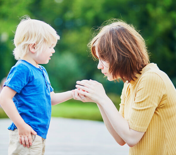 Mama kuca na ziemi, trzyma rękoma rączkę synka i dmucha w rączkę. Chłopczyk stoi i patrzy na mamę. Ma blond włosy, granatową koszulkę i beżowe spodnie. Mama ma brązowe włosy sięgające ramion i pomarańczową bluzkę z krótkim rękawem. W tle widać zieleń. 