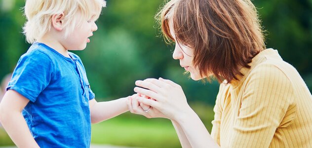 Mama kuca na ziemi, trzyma rękoma rączkę synka i dmucha w rączkę. Chłopczyk stoi i patrzy na mamę. Ma blond włosy, granatową koszulkę i beżowe spodnie. Mama ma brązowe włosy sięgające ramion i pomarańczową bluzkę z krótkim rękawem. W tle widać zieleń. 