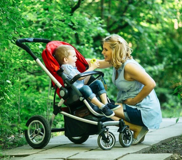 Mama i synek są w parku. Mama kuca obok synka, które siedzi w czerwonym wózku. Uśmiecha się do niego. Mama ma blond włosy, niebieską bluzkę i dżinsy przed kolano. Dziecko ma blond włosy, koszulkę i dżinsu. W tle widać zieleń. 