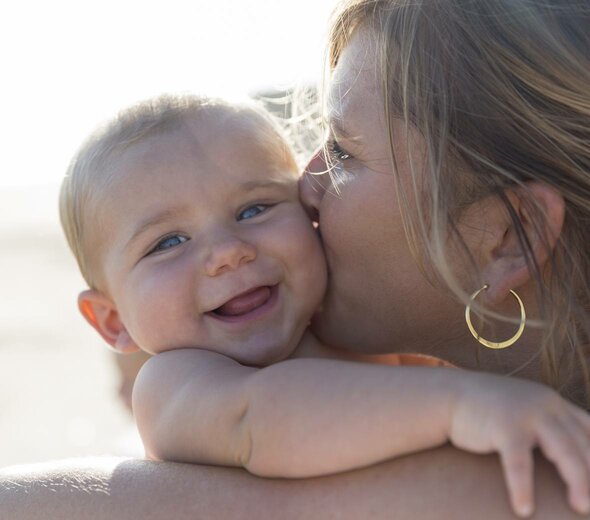 Mama trzyma na rękach niemowlę w słoneczny piękny dzień. Całuje je w główkę. Niemowlę patrzy w obiektyw i się śmieje. Mama ma blond włosy.