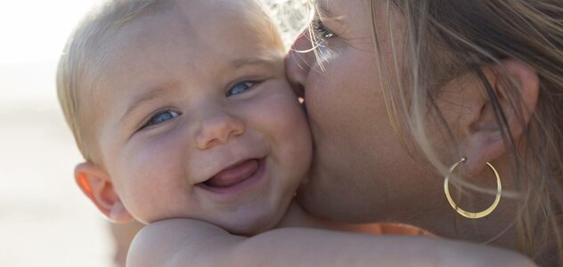 Mama trzyma na rękach niemowlę w słoneczny piękny dzień. Całuje je w główkę. Niemowlę patrzy w obiektyw i się śmieje. Mama ma blond włosy.