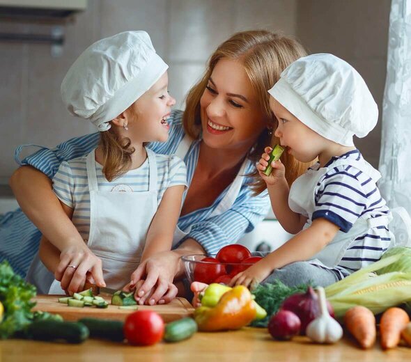 Mama i dwóje dzieci są w kuchni. Dzieci siedzą na blacie kuchennym wśród warzyw. Mają na głowach czapki kucharskie. Patrzą na mamę która się do nich śmieje. Chłopiec trzyma w rączce ogórka. Dziewczynka kroi wraz z mamą ogórka na drewnianej desce. 