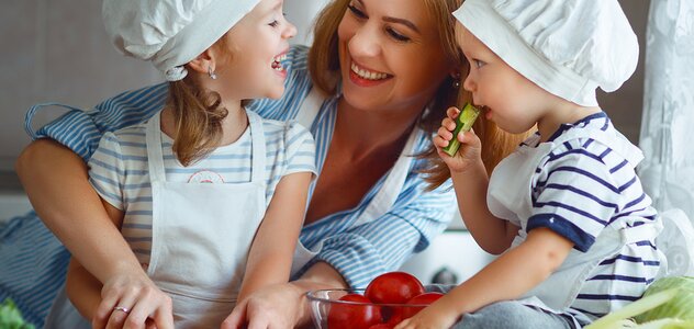 Mama i dwóje dzieci są w kuchni. Dzieci siedzą na blacie kuchennym wśród warzyw. Mają na głowach czapki kucharskie. Patrzą na mamę która się do nich śmieje. Chłopiec trzyma w rączce ogórka. Dziewczynka kroi wraz z mamą ogórka na drewnianej desce. 