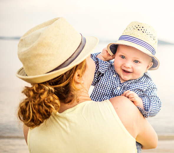 Mama trzyma na rękach niemowlę. Ma kapelusz letni. Niemowlę również ma na głowie kapelusz. Mama ma długie kręcone upięte włosy i żółtą koszulkę. Dziecko ma koszulę w biało granatową kratę. 