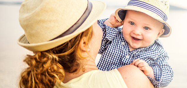 Mama trzyma na rękach niemowlę. Ma kapelusz letni. Niemowlę również ma na głowie kapelusz. Mama ma długie kręcone upięte włosy i żółtą koszulkę. Dziecko ma koszulę w biało granatową kratę. 