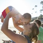 Mama siedzi na plaży nad morzem. Podnosi synka na rękach i całuje go w nosek. obok siedzi tata. Mama ma dżinsową niebieską bluzkę. Chłopczyk jest ubrany w kolorowe kąpielówki. 