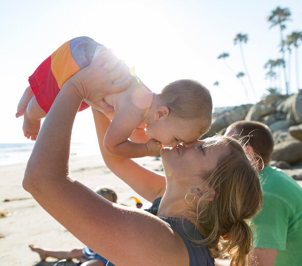 Mama siedzi na plaży nad morzem. Podnosi synka na rękach i całuje go w nosek. obok siedzi tata. Mama ma dżinsową niebieską bluzkę. Chłopczyk jest ubrany w kolorowe kąpielówki. 