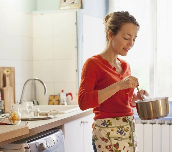 Kobieta stoi w kuchni i miesza drewnianą łyżką jedzenie w rondelku. Ma blond włosy, czerwony sweter i fartuch kuchenny przepasany w pasie. W tle widać okno, lodówkę, zmywarkę i rzeczy na blacie kuchennym. 