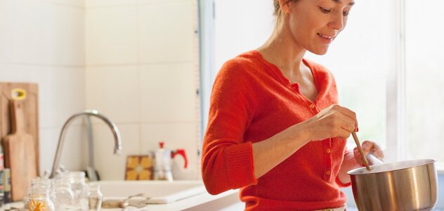 Kobieta stoi w kuchni i miesza drewnianą łyżką jedzenie w rondelku. Ma blond włosy, czerwony sweter i fartuch kuchenny przepasany w pasie. W tle widać okno, lodówkę, zmywarkę i rzeczy na blacie kuchennym. 