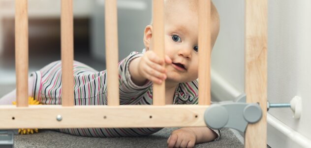 Niemowlę leży na brzuszku obok zamkniętej drewnianej bramki bezpieczeństwa przy schodach. Rączką łapie za szczebelek. Ma białe śpioszki w paski. Na podłodze leży szara wykładzina. Obok dziecka leży żółta piłeczka 