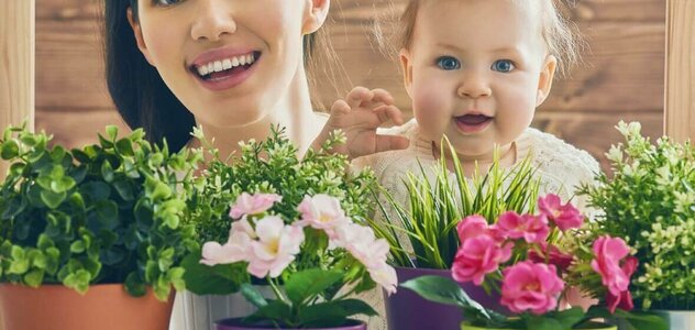 Mama i dziecko patrzą w obiektyw i uśmiechają się. Przed nimi stoi kilka rośli doniczkowych. Mama ma ciemne długie włosy. Dziecko ma na czubku głowy mały kucyk. W tle widać drewnianą ścianę. 