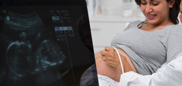 Kobieta w ciąży leży na łóżku w gabinecie lekarskim i podpiera się łokciami. Patrzy na swój brzuch. Lekarz przeprowadza USG płodu. Widać obraz płodu na monitorze. 