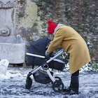 Mama stoi na chodniku w zimę obok wózka. Nachyla się nad wózkiem. Ma czerwoną czapkę, czarne kalosze i beżowy płaszcz. 