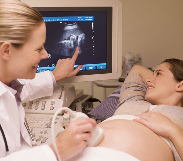 Kobieta leży w gabinecie lekarskim na łóżku. Lekarka przeprowadza badanie USG płodu. Uśmiecha się do kobiety w ciąży i pokazuje palcem obraz płodu na monitorze. Kobieta w ciąży patrzy na ekran monitora i  się uśmiecha. 